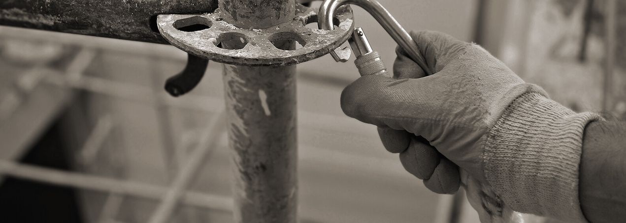 Eine behandschuhte Hand befestigt einen Karabiner an einem Metallgerüst und verdeutlicht damit die Sicherheitsvorkehrungen bei Bauarbeiten. Das Bild ist schwarz-weiß.