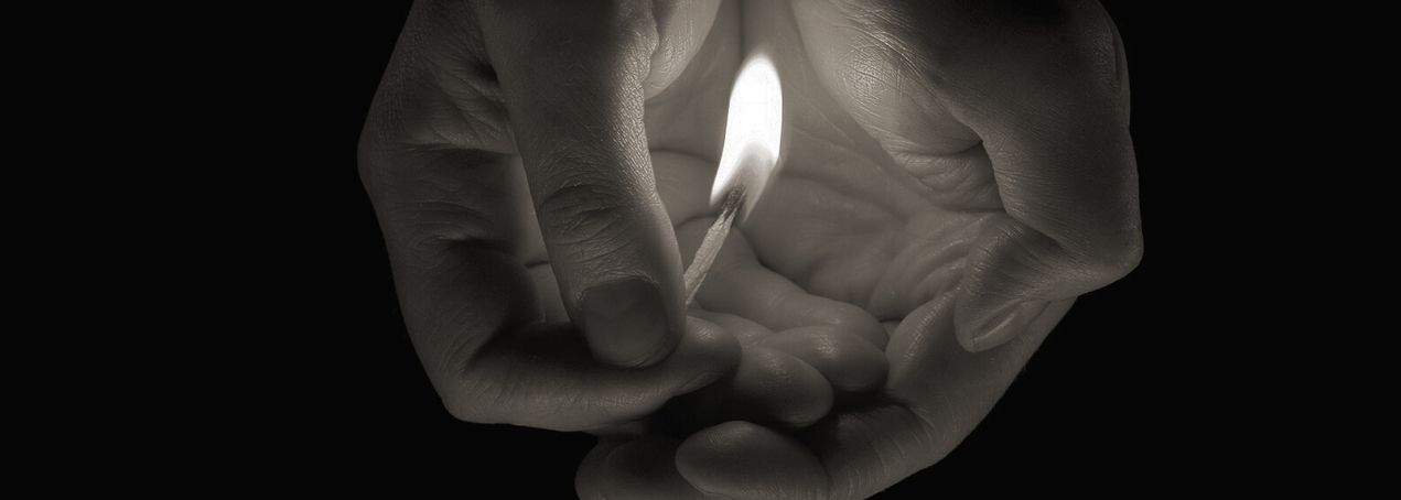 Zwei Hände halten sanft ein brennendes Streichholz und erhellen die Dunkelheit um sie herum. Der Fokus liegt auf der Flamme und den feinen Strukturen der Haut.