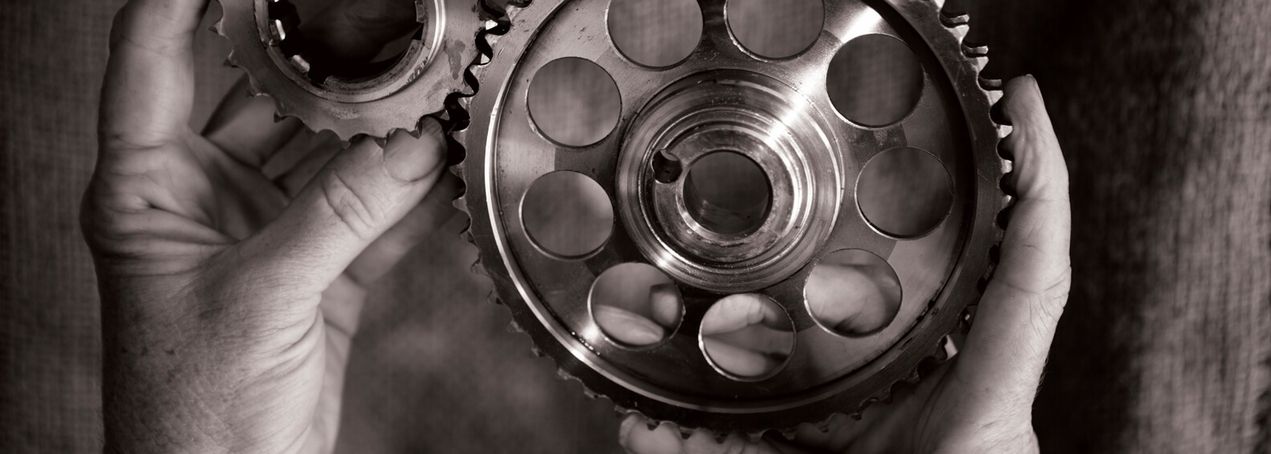 Zwei in Händen gehaltene Metallzahnräder mit aufwendigen Aussparungen und glänzenden Oberflächen heben sich von einem strukturierten Hintergrund ab.