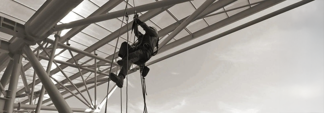 Person in Schutzkleidung und Helm klettert an Seilen an einem Stahlgerüst mit Querstreben und Trägern hoch.